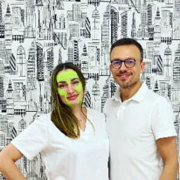 Kobieta z zielonymi taśmami kinesiotaping na twarzy, pozująca z mężczyzną na tle tapety w czarno-białe budynki.