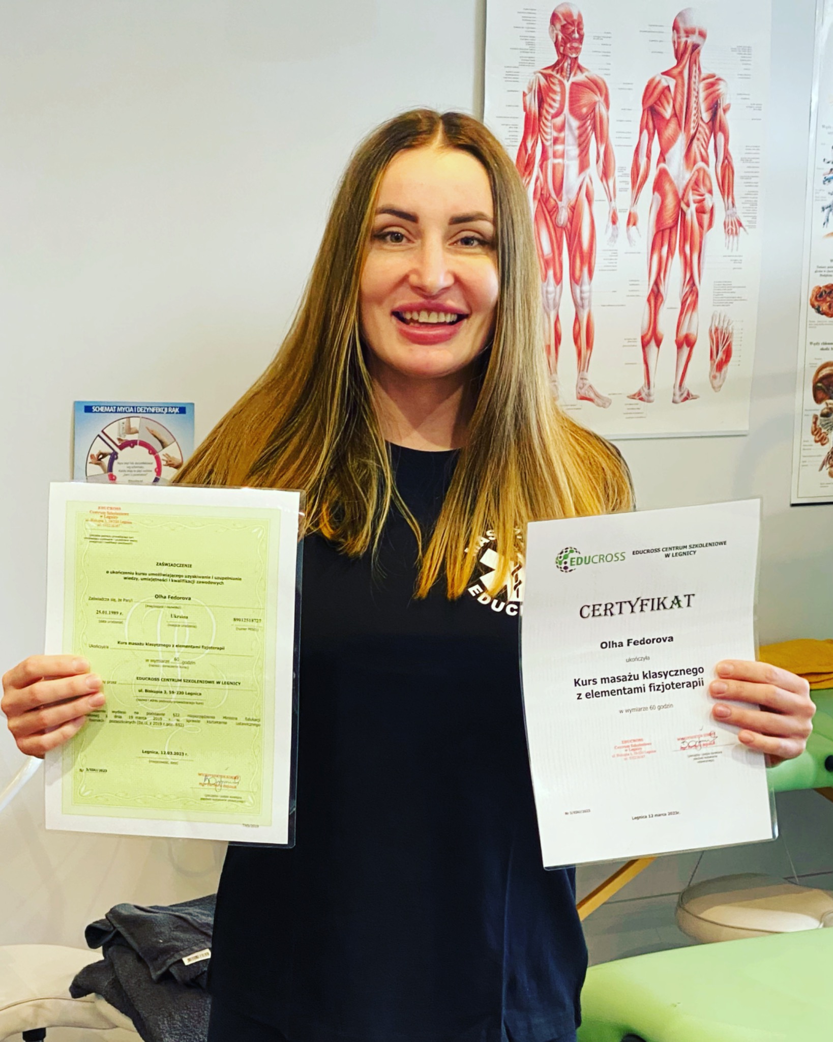 Uśmiechnięta kobieta prezentuje dwa certyfikaty ukończenia kursu masażu klasycznego z elementami fizjoterapii na tle plakatu z układem mięśniowym.