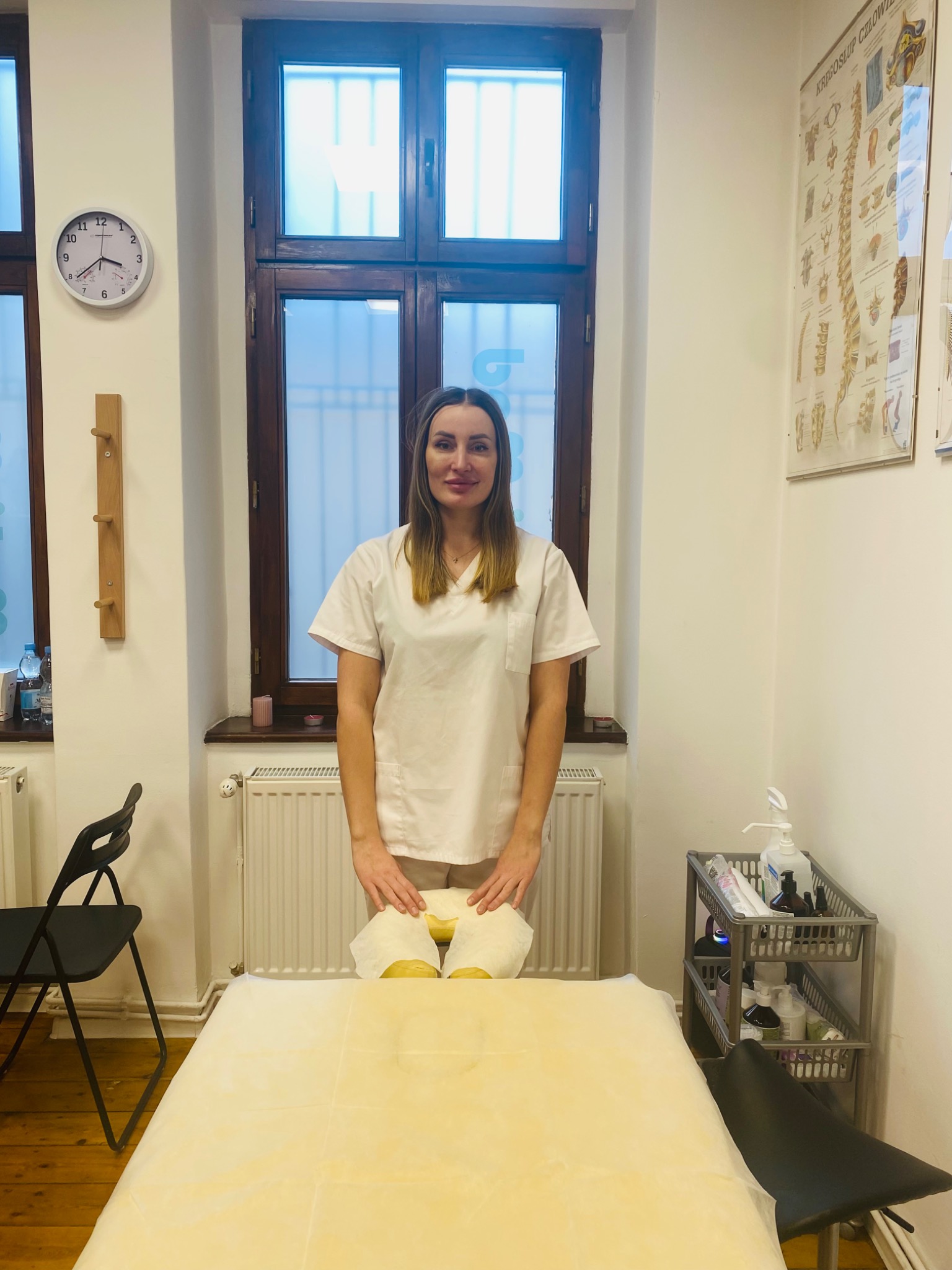 Kobieta w białym uniformie stoi za przygotowanym do zabiegu łóżkiem do masażu, widoczne akcesoria w tle i plakat z kręgosłupem na ścianie.