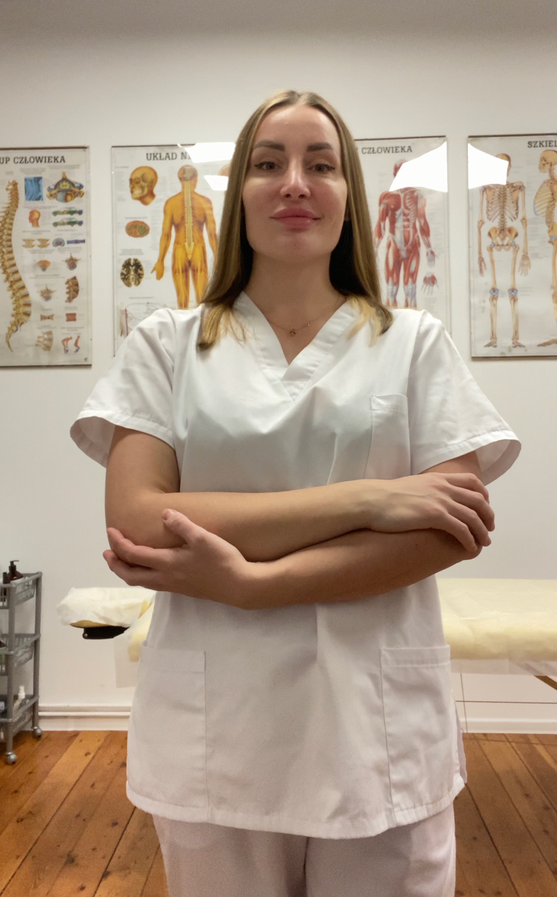 Młoda kobieta w białym uniformie medycznym, ze skrzyżowanymi rękami, na tle gabinetu z plakatami anatomicznymi i leżanką.