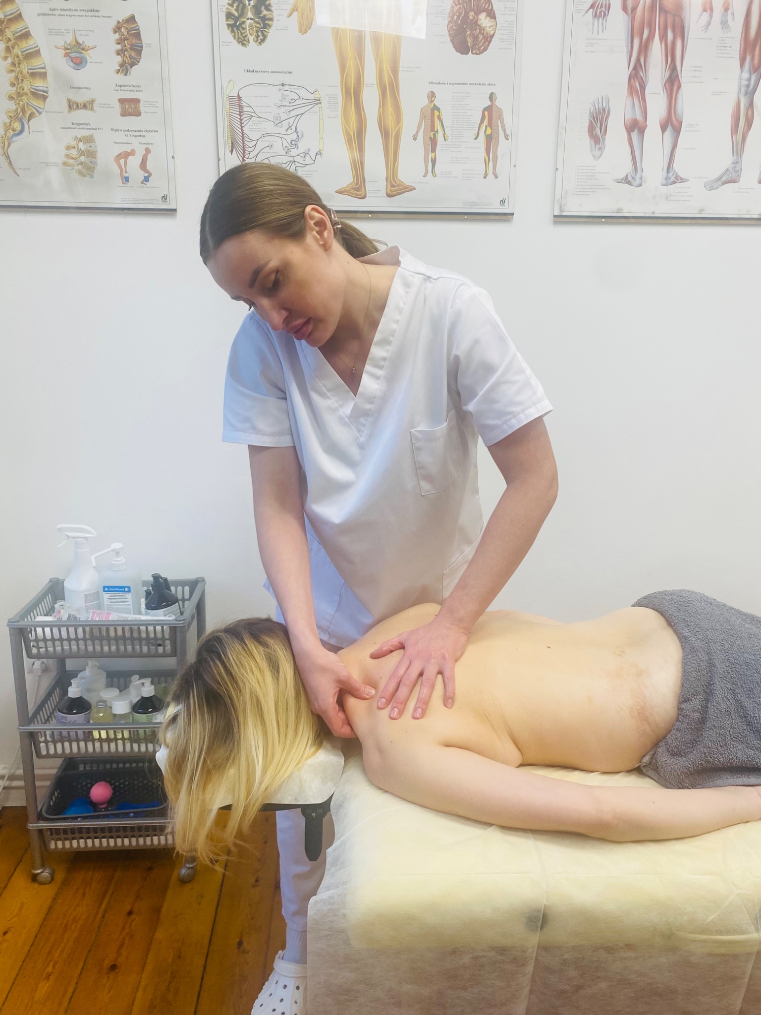 Masaż pleców i szyi wykonywany przez masażystkę w białym uniformie, widoczne plakaty anatomiczne na ścianie i wózek z akcesoriami do masażu.