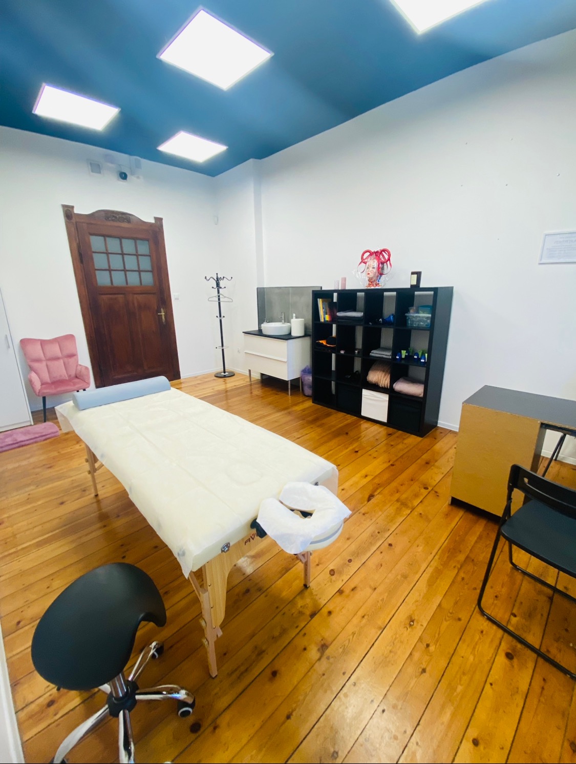 Jasne wnętrze gabinetu z drewnianą podłogą, centralnie ustawiony stół do masażu z pokrowcem, obok czarne krzesło na kółkach, w tle regał, umywalka i ozdobne dekoracje.