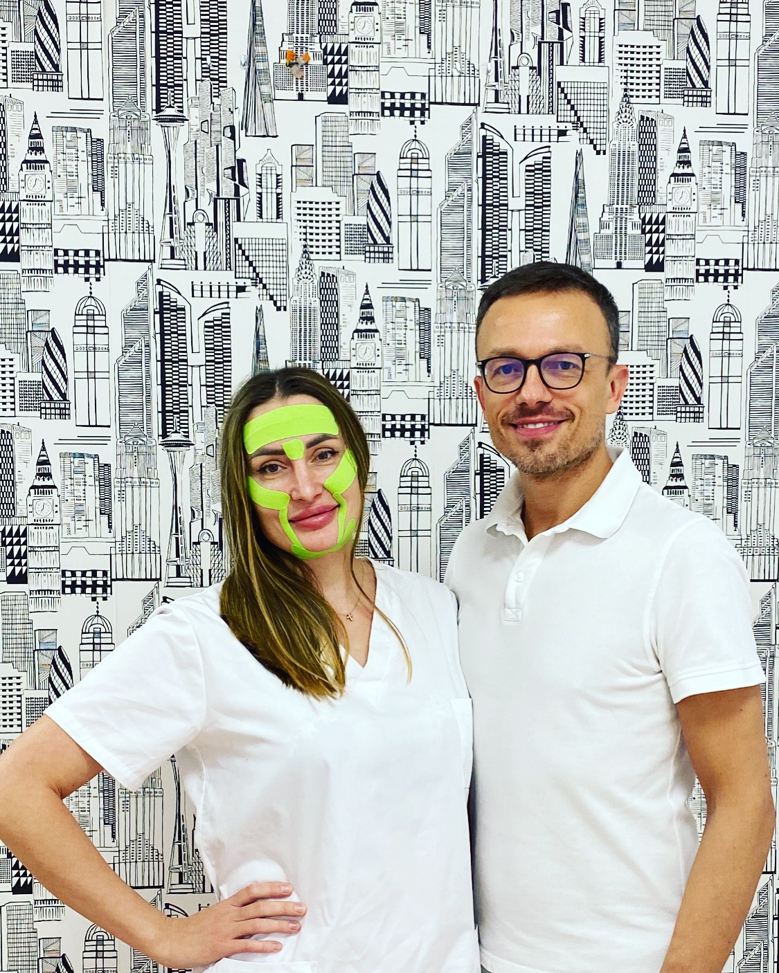 Kobieta z zielonymi taśmami kinesiotaping na twarzy, pozująca z mężczyzną na tle tapety w czarno-białe budynki.