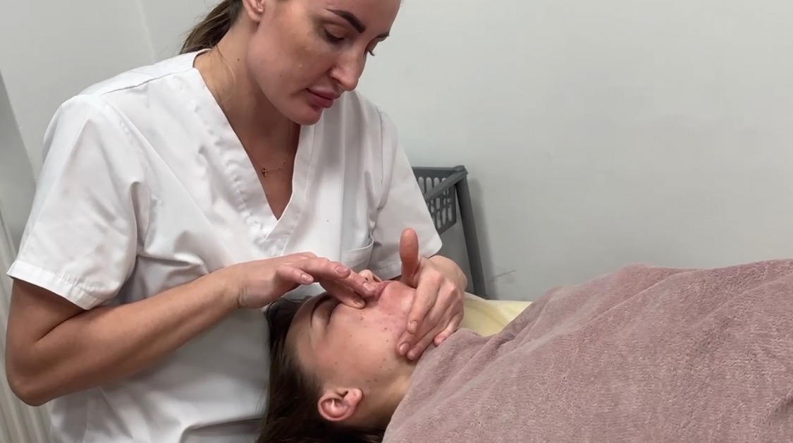 Kobieta w białym uniformie wykonuje masaż twarzy klientki leżącej na leżance, skupiając się na obszarze nosa i policzków.