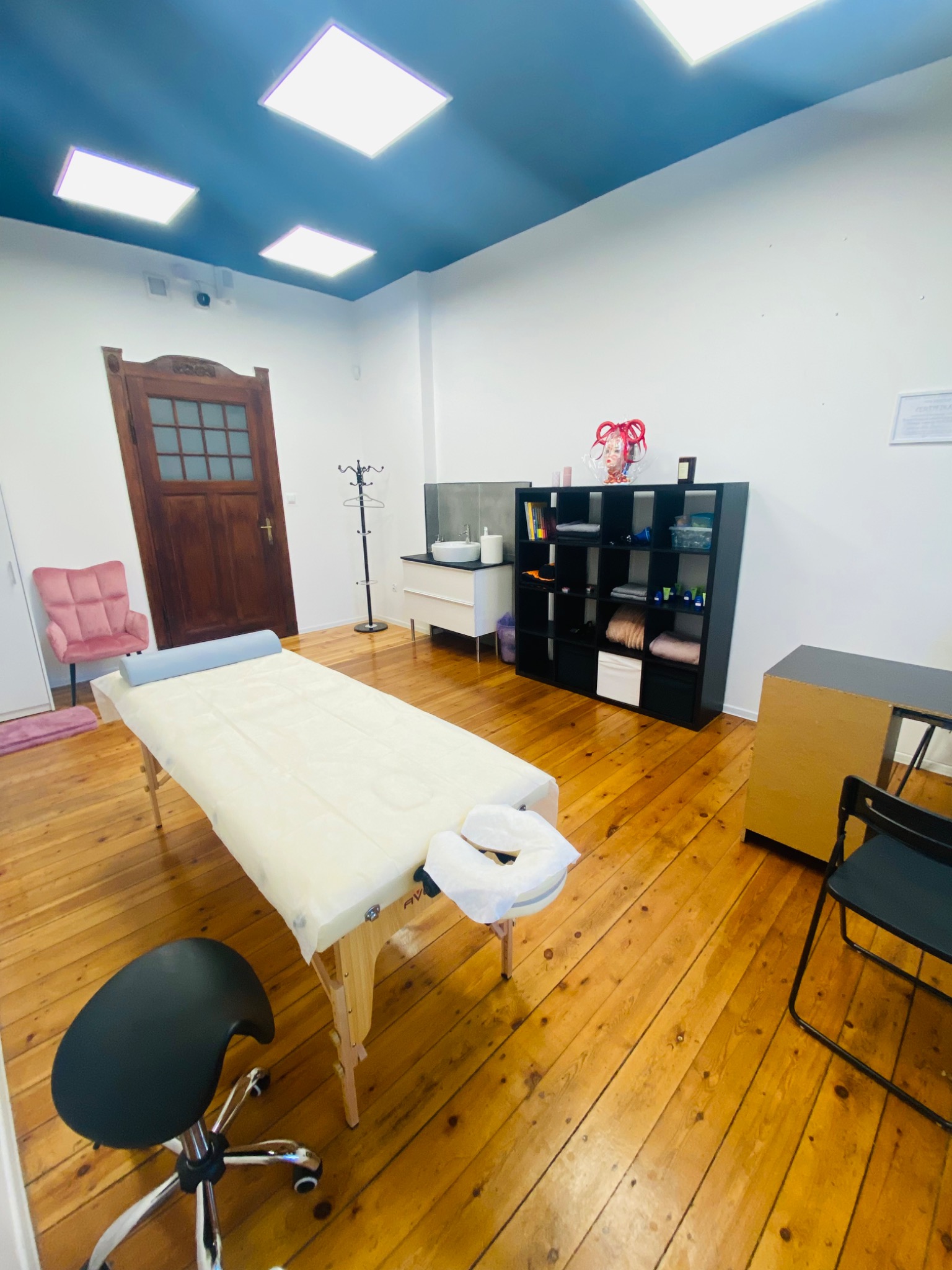 Jasne wnętrze gabinetu z drewnianą podłogą, centralnie ustawiony stół do masażu z poduszką, obok krzesło na kółkach, w tle szafka z kosmetykami i dekoracyjna głowa manekina.