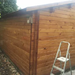 Świeżo pomalowana drewniana elewacja domku narzędziowego z budką dla ptaków, widoczna drabina malarska i otoczenie liściaste.