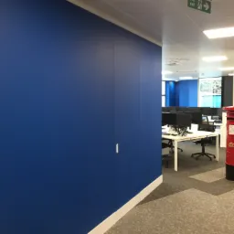 Świeżo pomalowana, intensywnie niebieska ściana w biurze, z widocznym fragmentem białej listwy przypodłogowej i dyskretnym uchwytem drzwiowym; w tle przestrzeń biurowa z biurkami i komputerami.