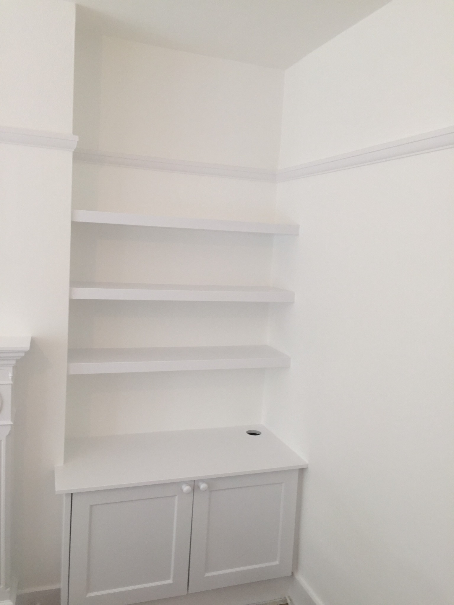 Wnęka z zabudową meblową i półkami, wszystko świeżo pomalowane na biało, widoczne detale wykończeniowe, takie jak listwy przypodłogowe i sztukateria.