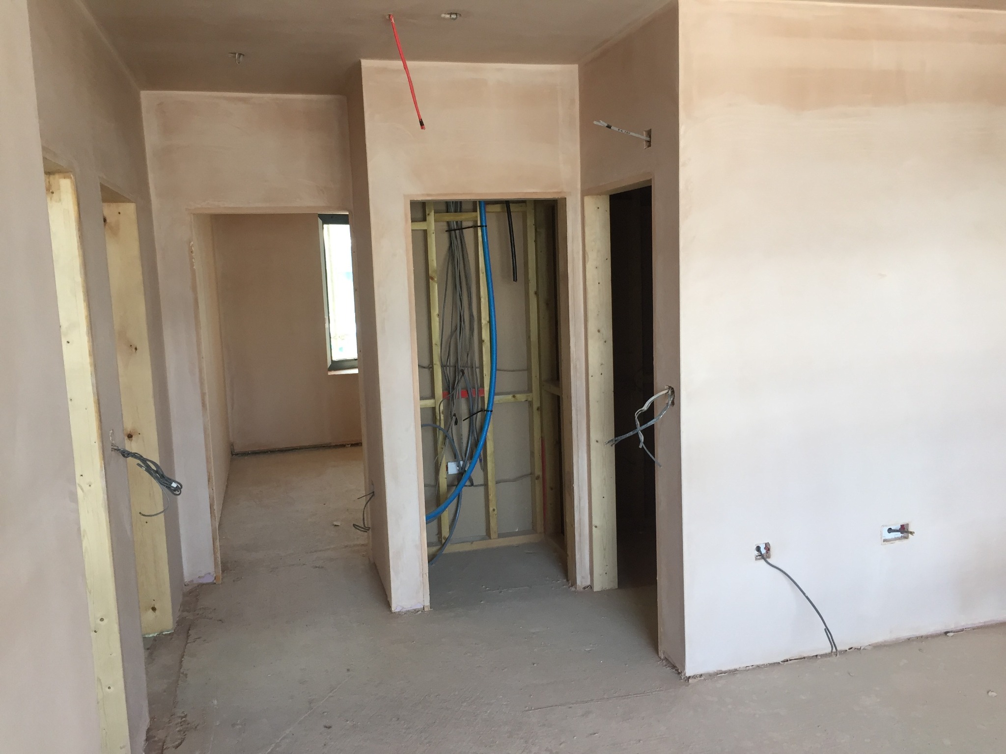 Wnętrze w trakcie prac wykończeniowych: ściany pokryte świeżo nałożoną warstwą szpachli, widoczne instalacje elektryczne i hydrauliczne w otworach ścian, surowy beton na podłodze.