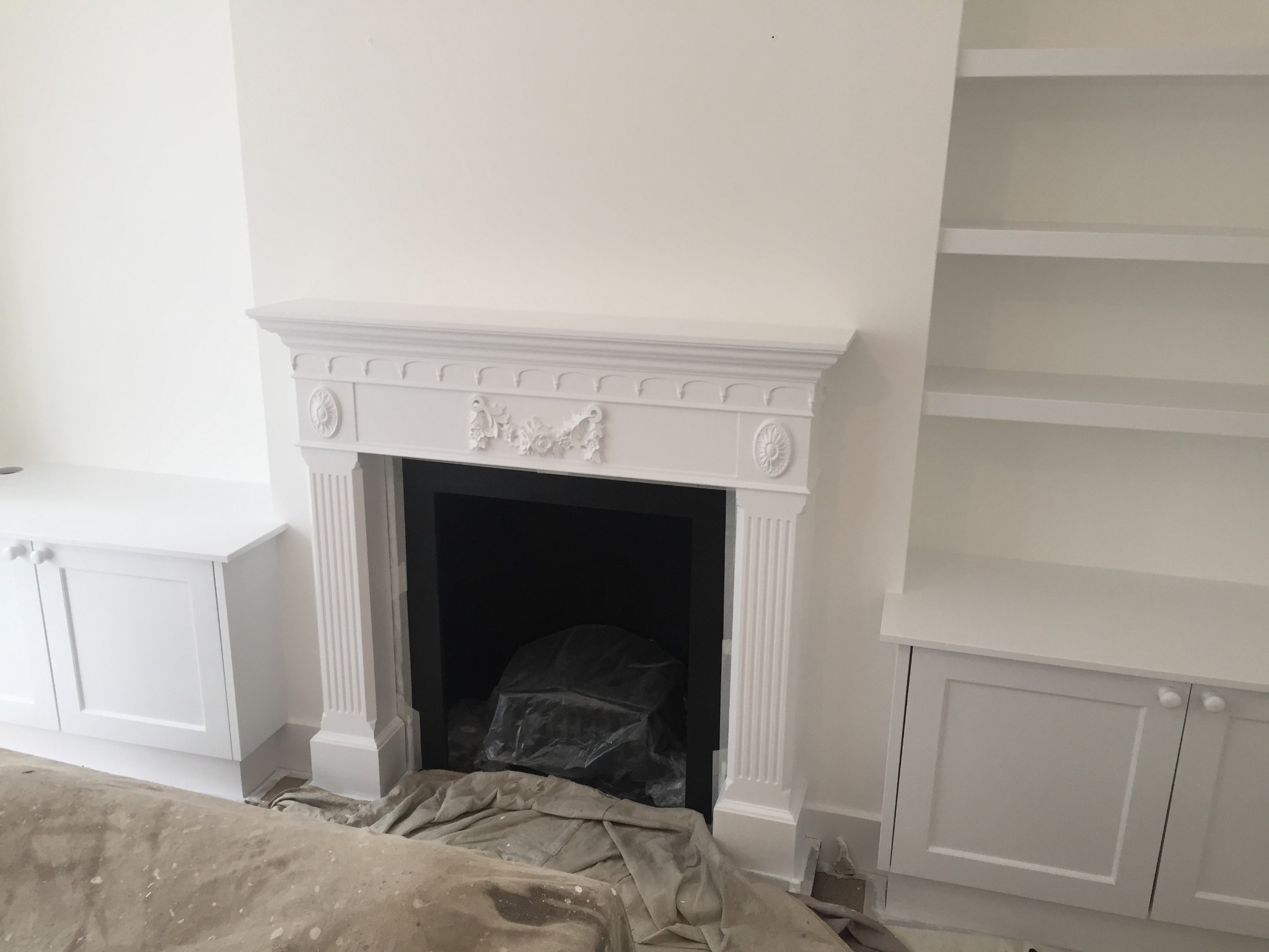 Białe wnętrze po odświeżeniu: pomalowane ściany, ozdobny kominek z zabezpieczeniem w środku, zabudowane szafki i półki, podłoga zabezpieczona folią malarską.