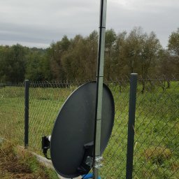 Łukasz Kołodziejczyk - ALSET-SYSTEM - Antena satelitarna zamontowana na metalowym słupku przy ogrodzeniu z siatki, z widoczną puszką antenową na górze słupka oraz niebieską rurką karbowaną osłaniającą przewody u dołu.