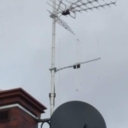 Łukasz Kołodziejczyk - ALSET-SYSTEM - Antena telewizyjna i satelitarna zamontowana na dachu budynku z ceglanym kominem, widok z dołu na tle szarego nieba.