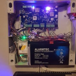 Łukasz Kołodziejczyk - ALSET-SYSTEM - Otwarta obudowa centrali alarmowej z widoczną płytą główną, akumulatorem marki Alarmtec i licznymi przewodami. Świecące diody LED na płycie wskazują na aktywność systemu.