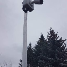 Łukasz Kołodziejczyk - ALSET-SYSTEM - Kamera przemysłowa zamontowana na metalowym słupie, skierowana w prawo, na tle pochmurnego nieba i drzew iglastych.