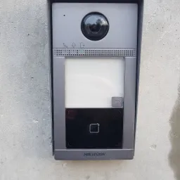 Szary wideodomofon Hikvision zamontowany na surowej, betonowej ścianie, widoczny obiektyw kamery, ekran, czytnik kart i podświetlany przycisk.