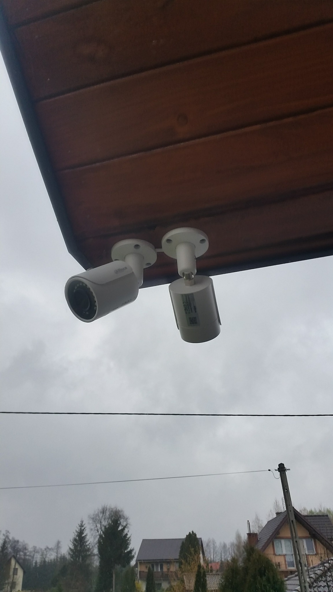 Dwie kamery cylindryczne marki Dahua zamontowane pod drewnianym okapem, widok z dołu na tle pochmurnego nieba i zabudowań mieszkalnych.