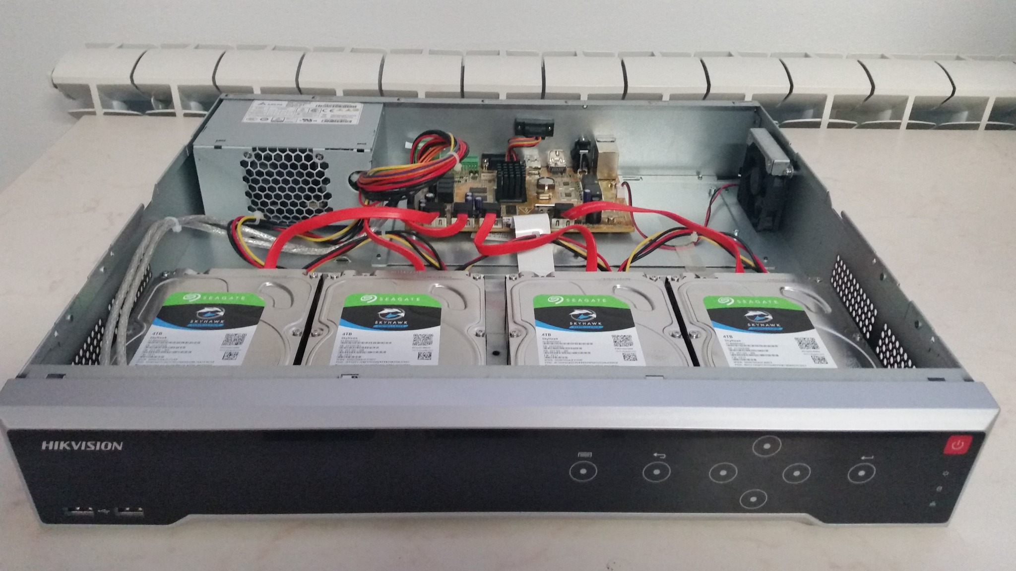 Otwarta obudowa rejestratora Hikvision z czterema dyskami twardymi Seagate Skyhawk 4TB, zasilaczem, płytą główną i wentylatorem, widoczne kable SATA i zasilające.