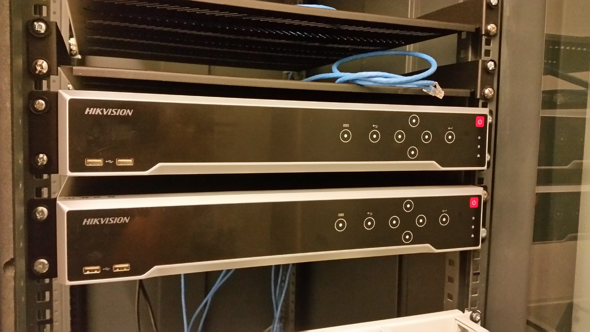Dwa rejestratory Hikvision zamontowane w szafie rack, widoczne porty USB, przyciski funkcyjne, diody LED i niebieski kabel sieciowy na górnej półce.