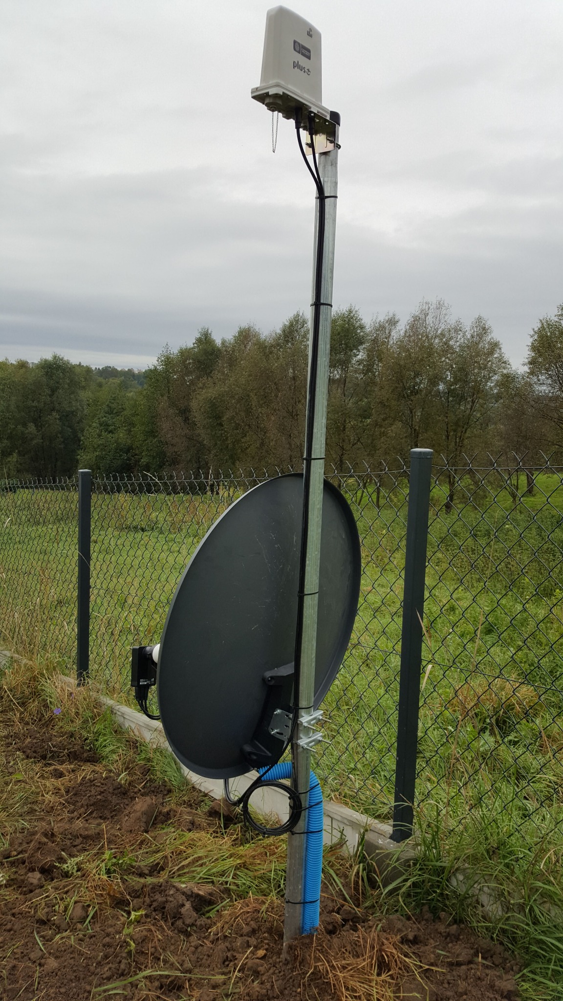 Antena satelitarna zamontowana na metalowym słupku przy ogrodzeniu z siatki, z widoczną puszką antenową na górze słupka oraz niebieską rurką karbowaną osłaniającą przewody u dołu.
