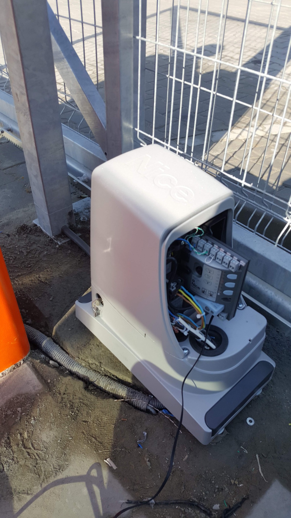 Otwarta obudowa automatyki bramy przesuwnej marki Nice, widoczne okablowanie i sterownik, zamontowana przy metalowym ogrodzeniu na nierównym podłożu.