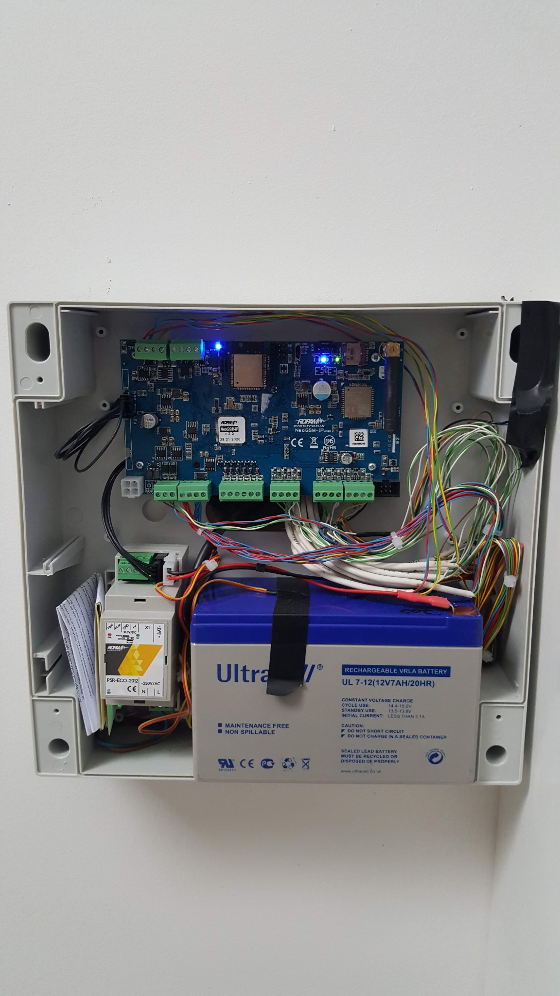Centrala alarmowa w szarej obudowie z widoczną płytką drukowaną, licznymi przewodami, transformatorem PSR-ECO-2012 i akumulatorem UltraCell UL 7-12. Dwie niebieskie diody LED świecą na płytce.
