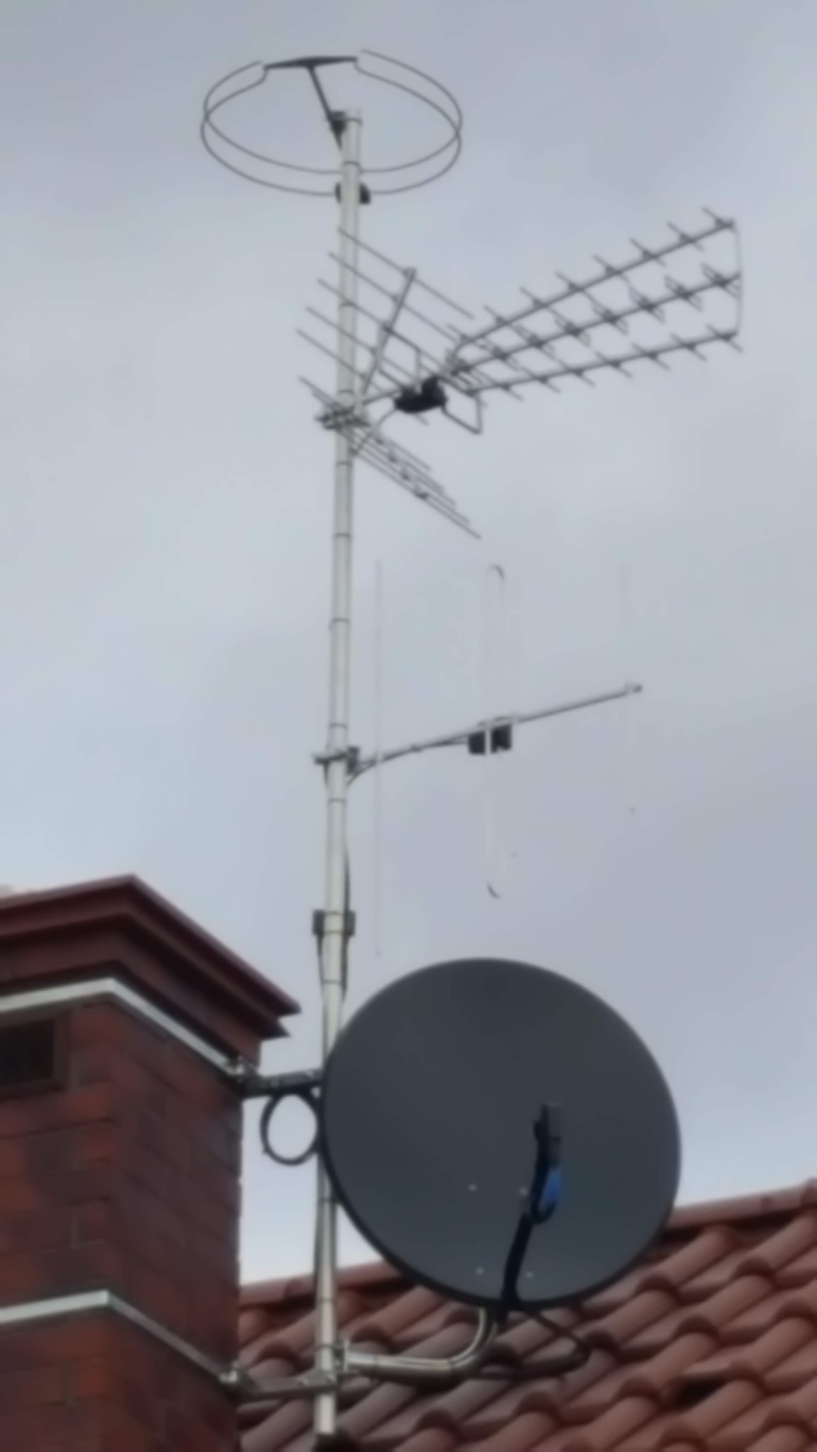 Antena telewizyjna i satelitarna zamontowana na dachu budynku z ceglanym kominem, widok z dołu na tle szarego nieba.
