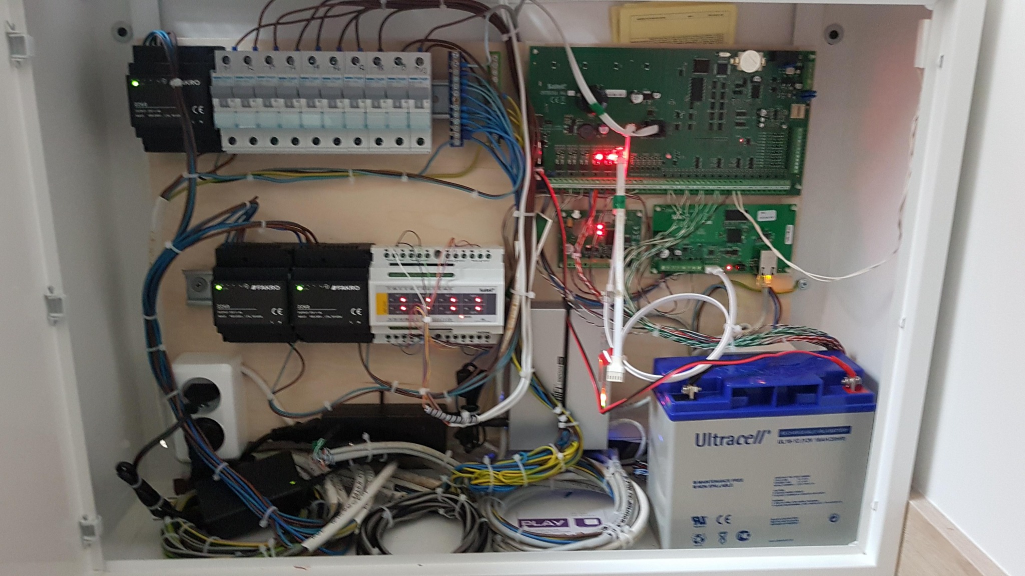 Otwarta szafka elektryczna z widocznymi elementami alarmu: płytki drukowane, przekaźniki Fakro, bezpieczniki, akumulator Ultracell, liczne przewody i złącza.
