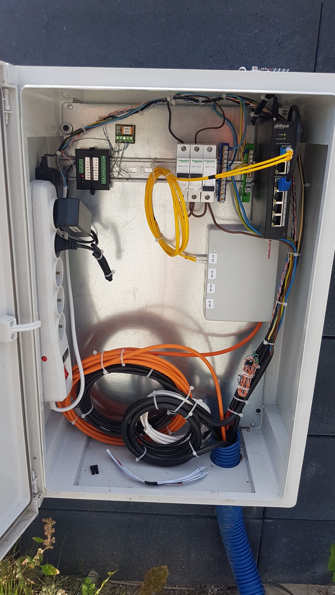 Otwarta skrzynka instalacyjna z widocznym okablowaniem, listwą zasilającą, modułem światłowodowym Ultimode, przełącznikiem sieciowym Dahua, bezpiecznikami Schneider Electric oraz innymi elementami...