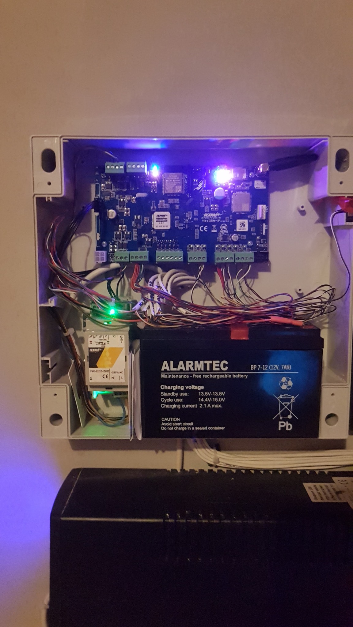 Otwarta obudowa centrali alarmowej z widoczną płytą główną, akumulatorem marki Alarmtec i licznymi przewodami. Świecące diody LED na płycie wskazują na aktywność systemu.