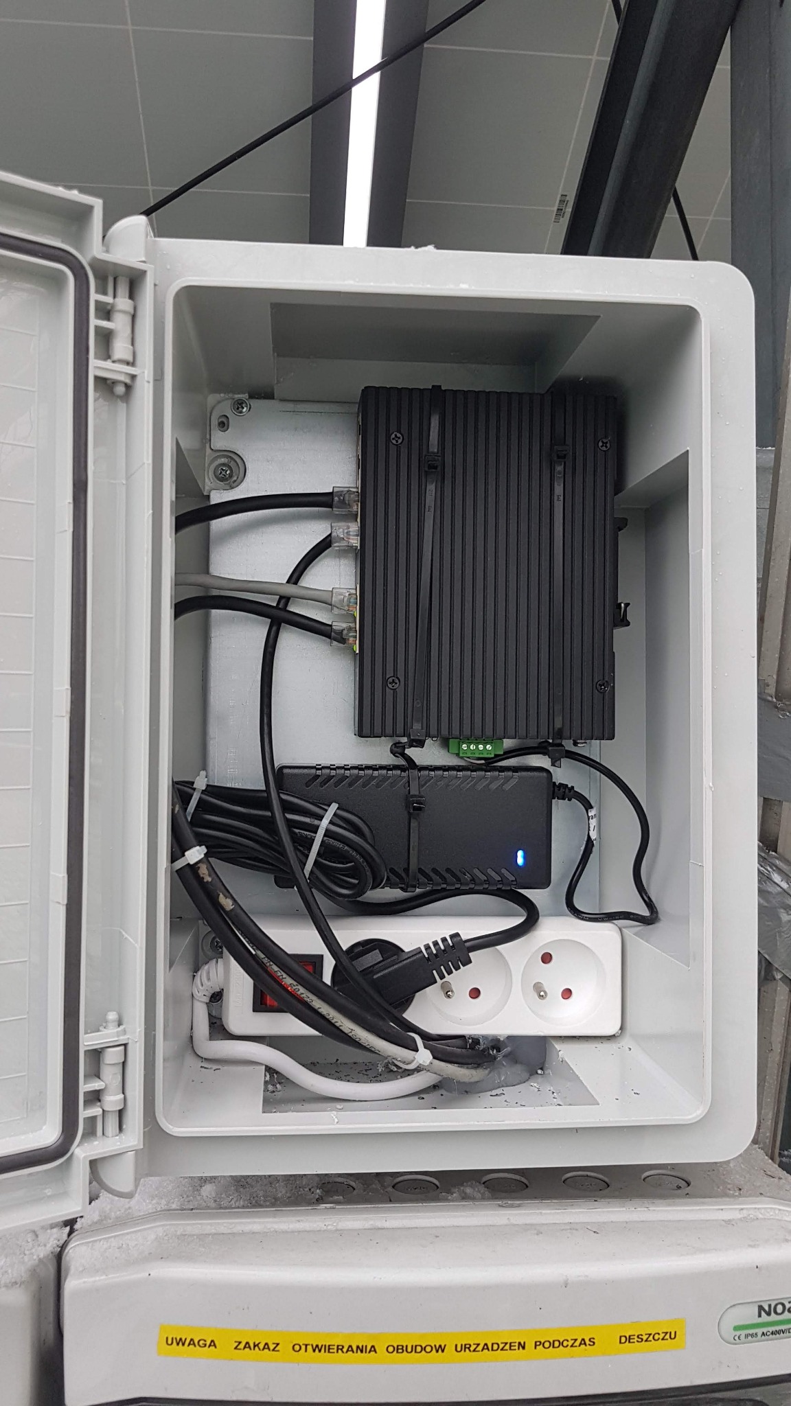 Wnętrze otwartej, białej skrzynki z urządzeniem sieciowym, zasilaczem z niebieską diodą, listwą zasilającą z włącznikiem i kablami. Widoczny napis ostrzegawczy o zakazie otwierania obudowy podczas...