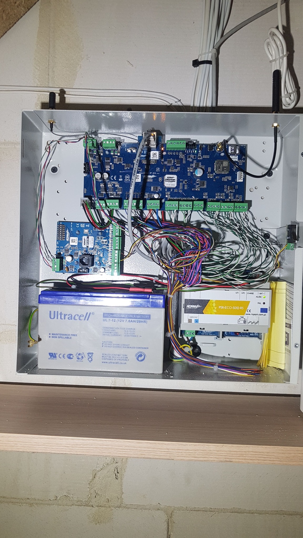 Otwarta szafka sterownicza z elektroniką, akumulatorem Ultracell i przewodami, zamontowana na ścianie z widoczną instalacją elektryczną i elementami systemu zasilania awaryjnego.