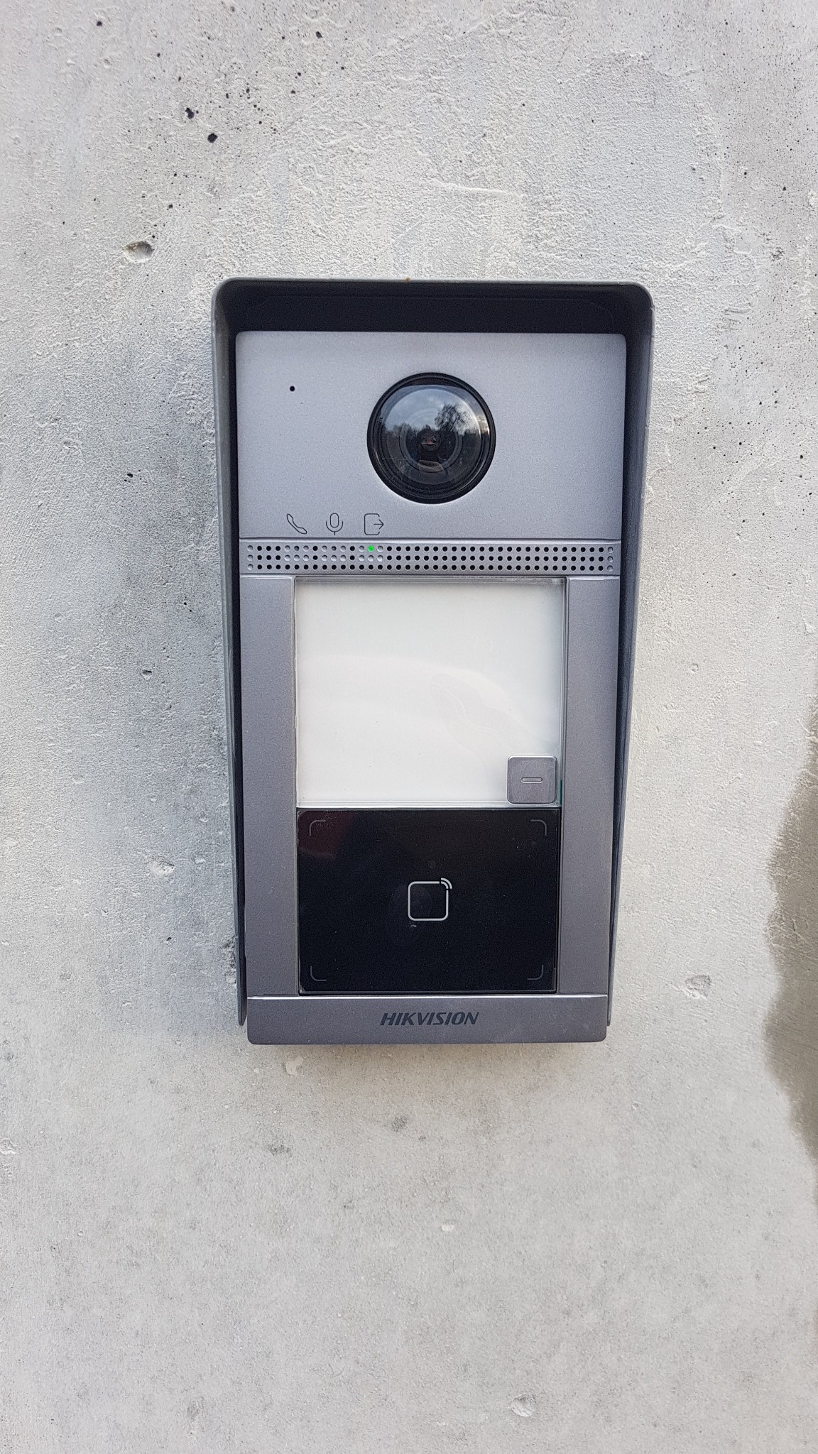 Szary wideodomofon Hikvision zamontowany na surowej, betonowej ścianie, widoczny obiektyw kamery, ekran, czytnik kart i podświetlany przycisk.