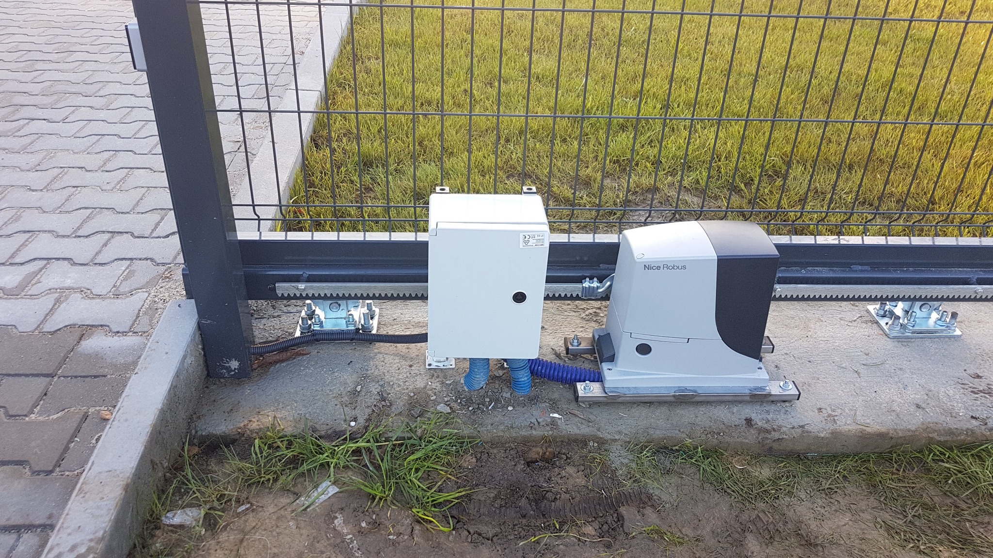 Zamontowany napęd do bramy przesuwnej Nice Robus z widoczną listwą zębatą i okablowaniem w niebieskiej rurce karbowanej.