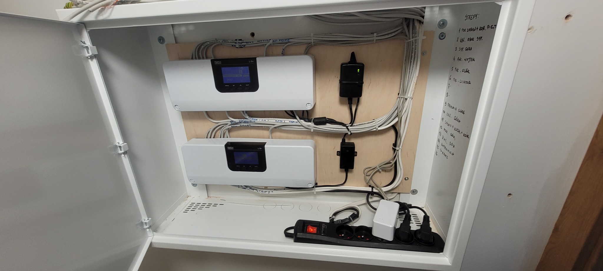 Szafka z dwoma białymi modułami sterującymi TECH L-9, okablowaniem, zasilaczami i listwą zasilającą; na drzwiach szafki odręcznie wypisane nazwy stref alarmowych.
