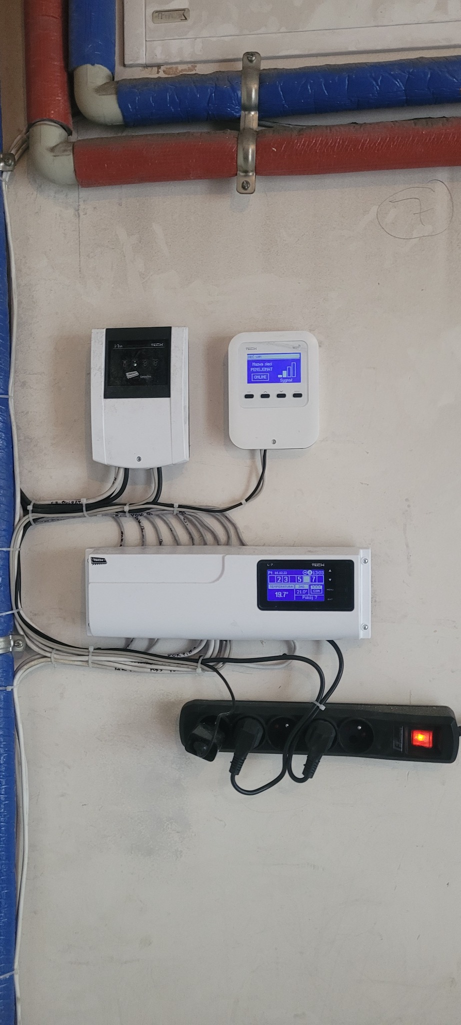 Elementy systemu inteligentnego domu zamontowane na ścianie w Krakowie, widoczne przewody, listwa zasilająca z włączonym zasilaniem oraz rury grzewcze. Na wyświetlaczach urządzeń widoczne parametry...