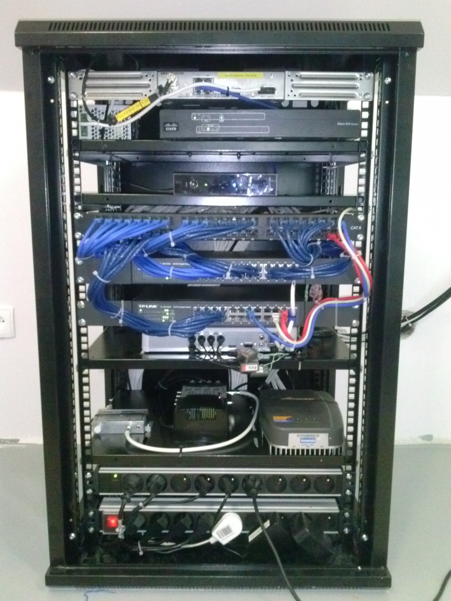 Szafa rack z zainstalowanymi urządzeniami sieciowymi: router Cisco, switche z niebieskimi kablami Ethernet, listwa zasilająca z włączonym czerwonym przyciskiem, zasilacz awaryjny UPS.