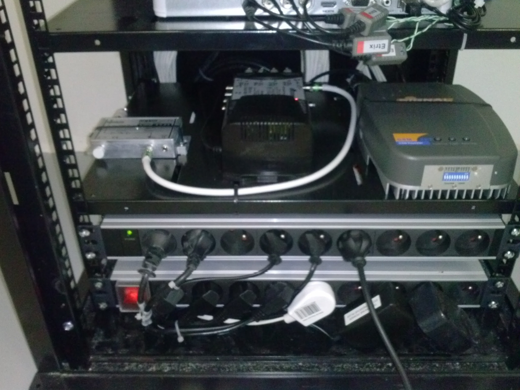 Szafa rack z zainstalowanym sprzętem: zasilacz awaryjny UPS, listwa zasilająca z wpiętymi kablami, oraz inne urządzenia elektroniczne z licznymi przewodami.