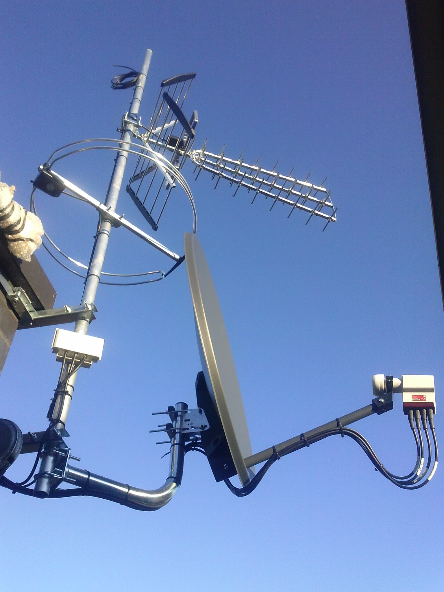 Zestaw antenowy: satelitarna, szerokopasmowa i Yagi-Uda zamontowane na jednym maszcie, widoczne z dołu na tle błękitnego nieba.
