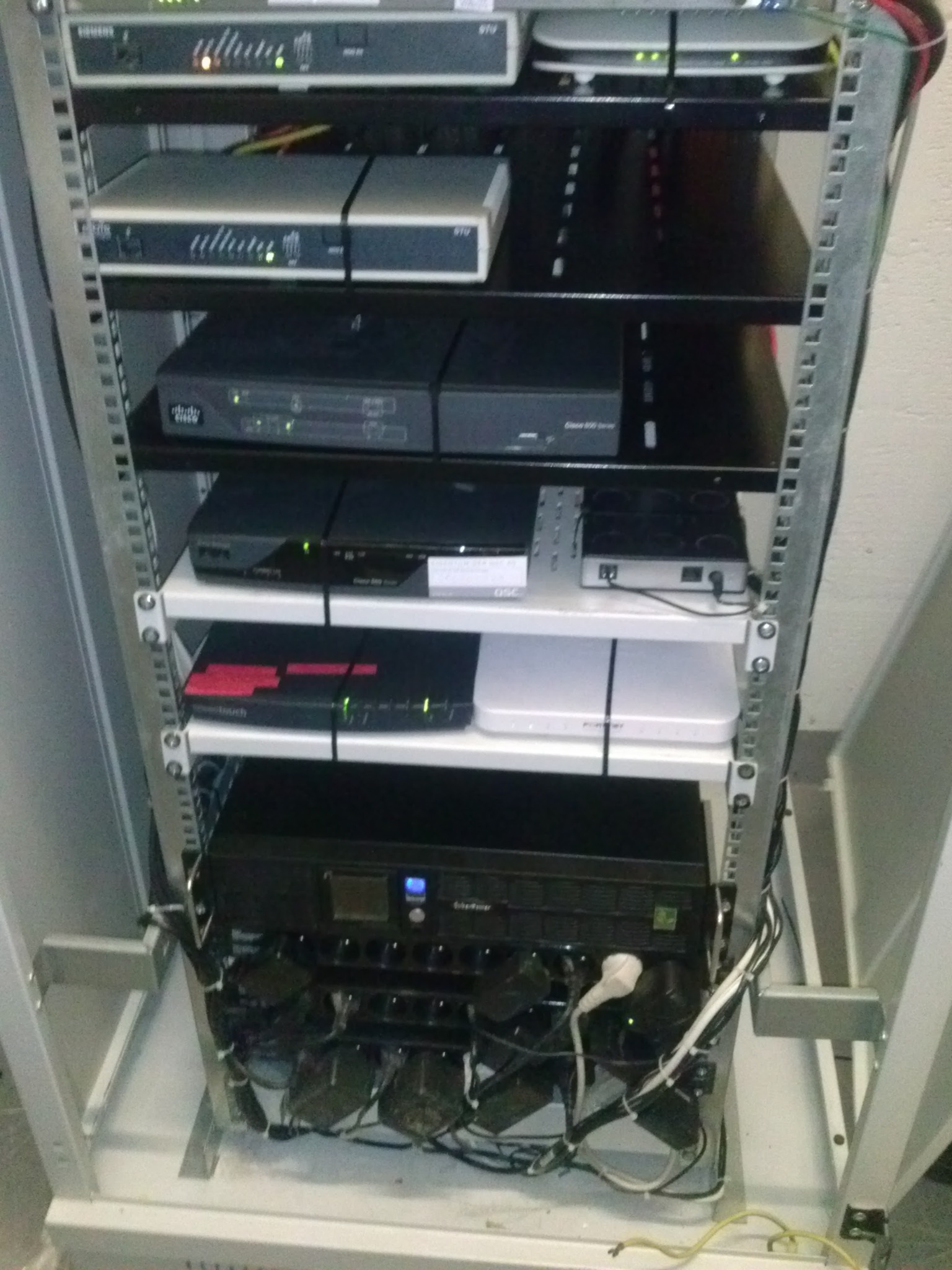 Regał serwerowy z różnymi urządzeniami sieciowymi, widoczne kable i zasilacze, część urządzeń sygnalizuje działanie diodami.