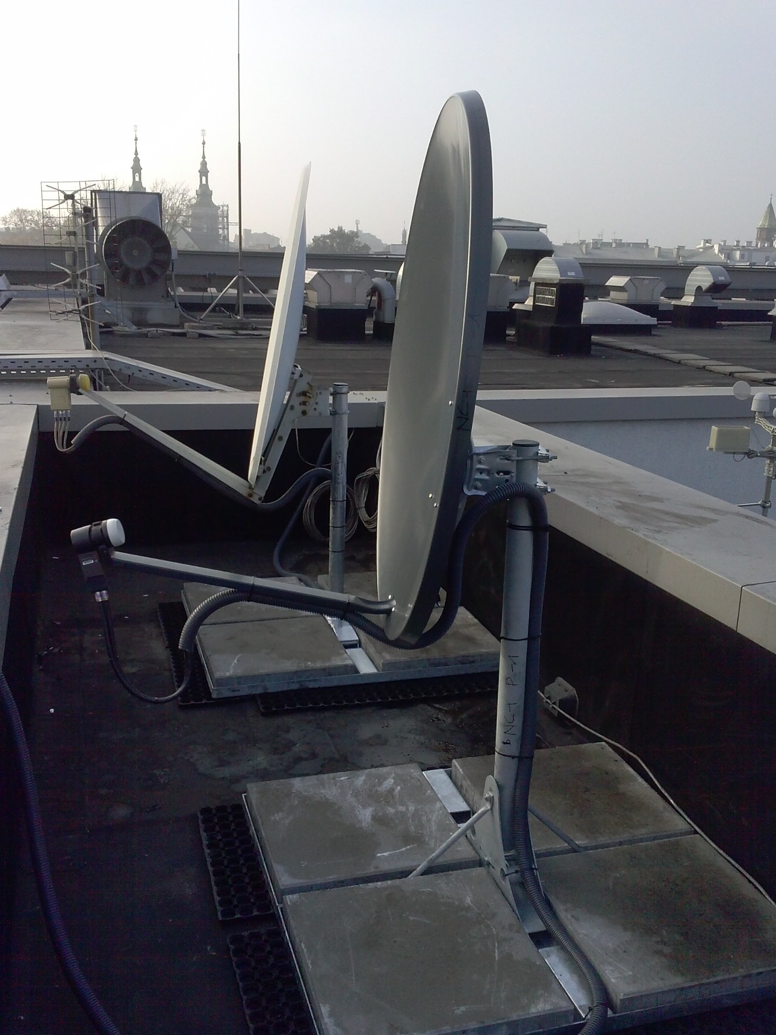 Dwie anteny satelitarne na dachu budynku, widoczne kable i mocowania, w tle zabudowa miejska z wieżami kościołów w oddali, pochmurne niebo.