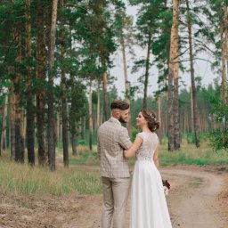 Kristina Maciejewska - Para młoda, on w beżowym garniturze w kratę, ona w białej sukni z koronkową górą, stoją na leśnej drodze, patrząc na siebie z uśmiechem.
