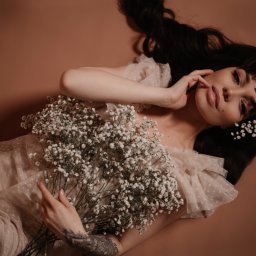 Kristina Maciejewska - Kobieta z ciemnymi włosami leży na beżowym tle, trzymając bukiet gipsówki i mając pojedyncze kwiaty we włosach. Widoczny tatuaż na ramieniu.