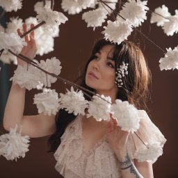 Kristina Maciejewska - Portret kobiety w zwiewnej sukience, otoczonej gałązkami z białymi, papierowymi kwiatami; delikatny makijaż i kwiaty we włosach; tatuaż na przedramieniu.