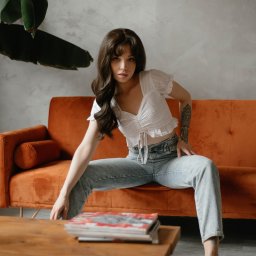 Kristina Maciejewska - Portret kobiety siedzącej na pomarańczowej sofie, ubrana w jeansy i białą bluzkę, z tatuażem na ramieniu, pozującej w studio z drewnianym stolikiem i magazynami.
