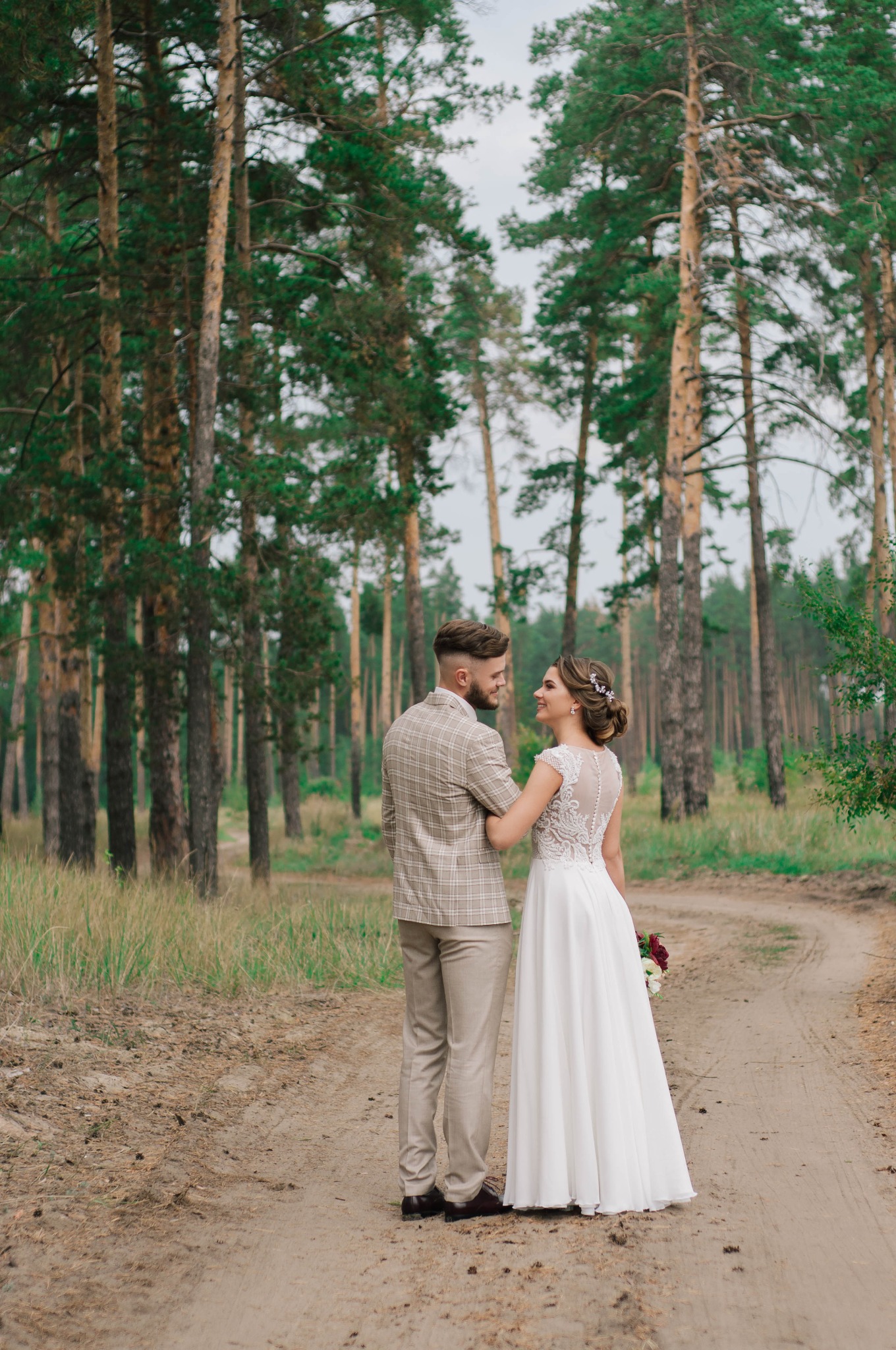 Para młoda, on w beżowym garniturze w kratę, ona w białej sukni z koronkową górą, stoją na leśnej drodze, patrząc na siebie z uśmiechem.