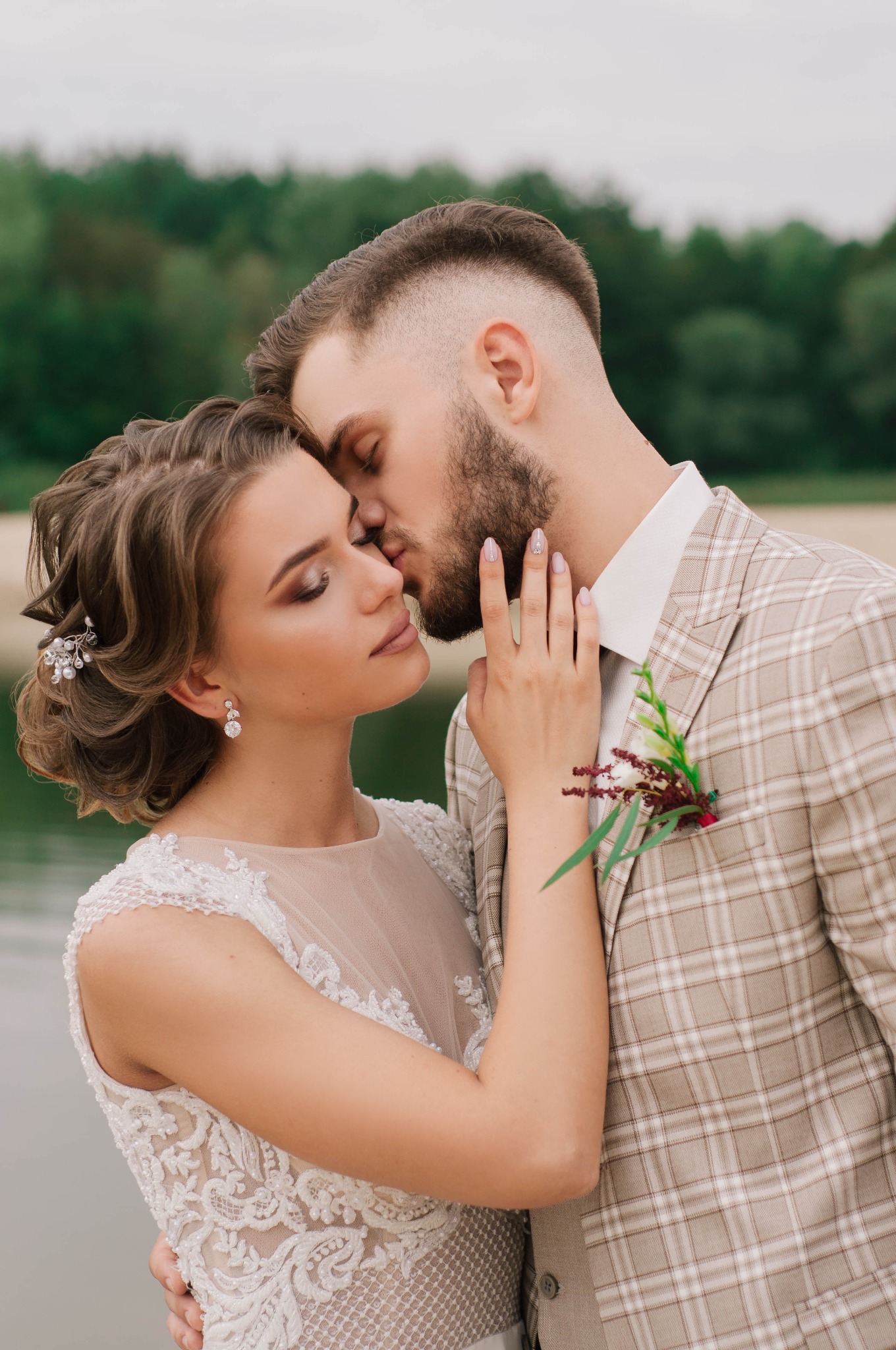 Para młoda w objęciach: panna młoda w koronkowej sukni i z upiętymi włosami, pan młody w kraciastej marynarce, delikatny pocałunek na tle zieleni i wody.