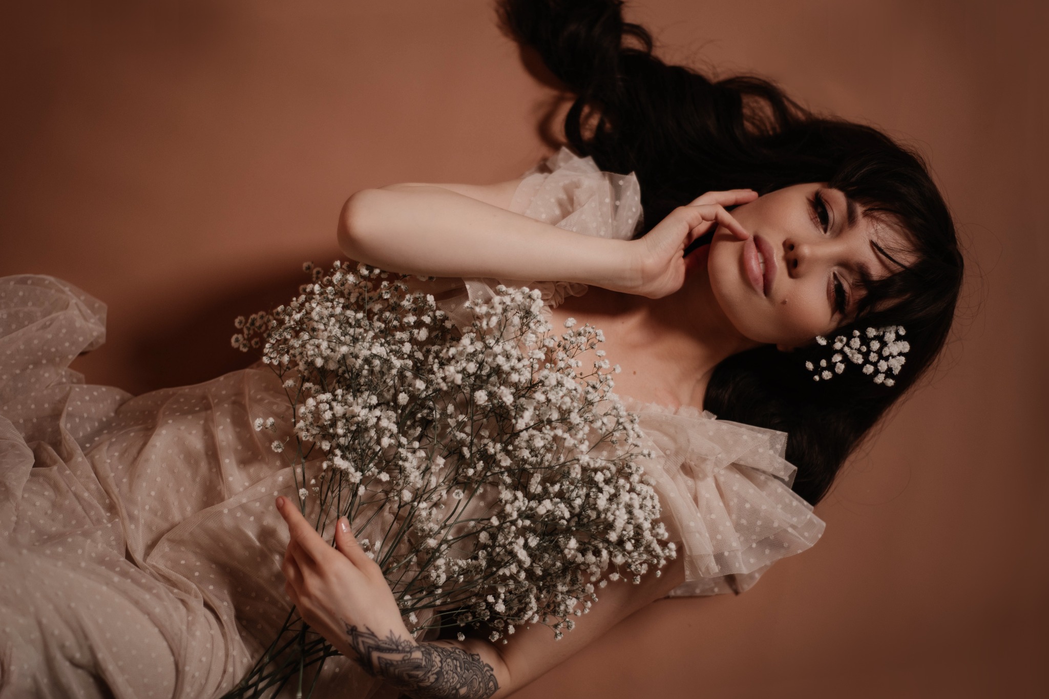 Kobieta z ciemnymi włosami leży na beżowym tle, trzymając bukiet gipsówki i mając pojedyncze kwiaty we włosach. Widoczny tatuaż na ramieniu.