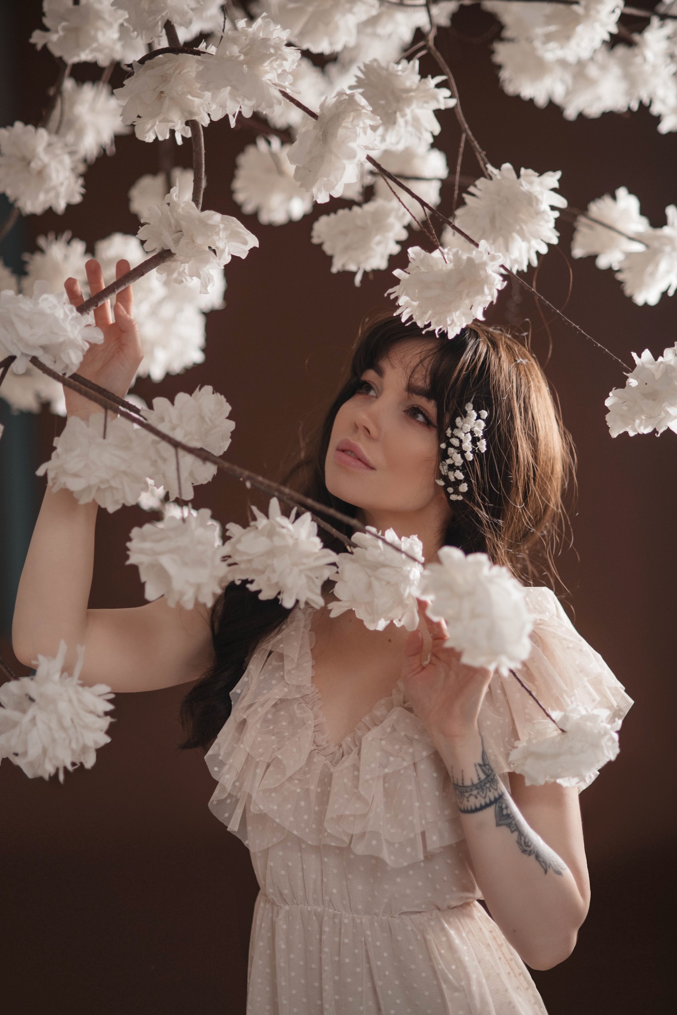 Portret kobiety w zwiewnej sukience, otoczonej gałązkami z białymi, papierowymi kwiatami; delikatny makijaż i kwiaty we włosach; tatuaż na przedramieniu.