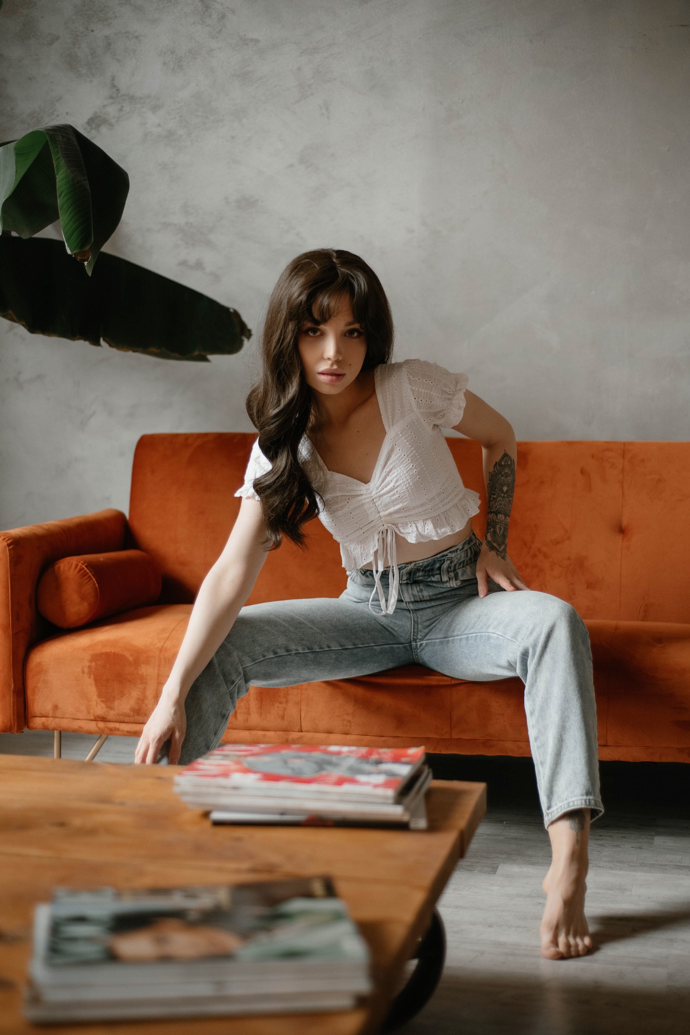 Portret kobiety siedzącej na pomarańczowej sofie, ubrana w jeansy i białą bluzkę, z tatuażem na ramieniu, pozującej w studio z drewnianym stolikiem i magazynami.