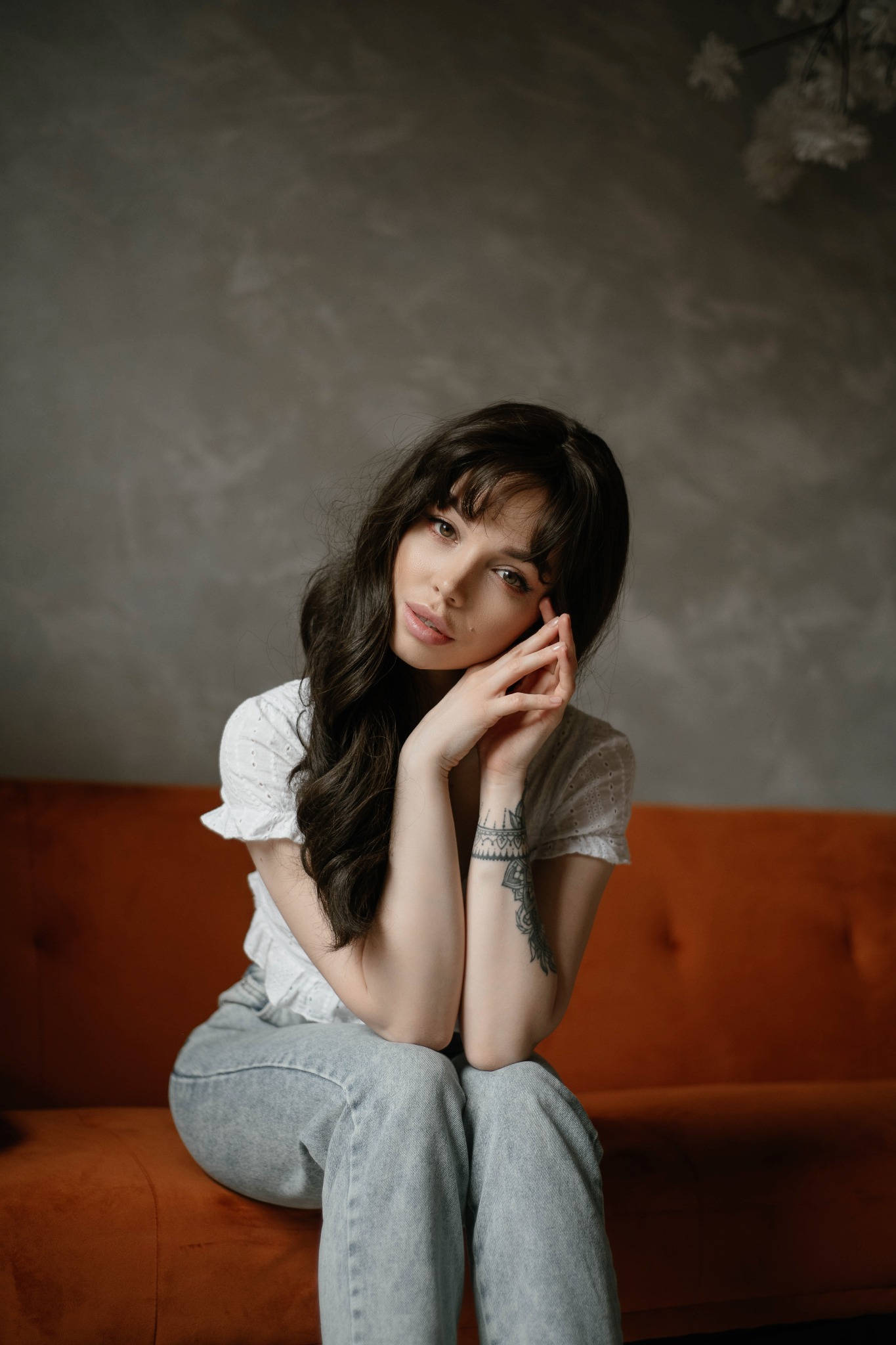 Portret kobiety z ciemnymi, falowanymi włosami, opierającej brodę na splecionych dłoniach, siedzącej na pomarańczowej sofie na tle szarej ściany z dekoracyjną gałązką.